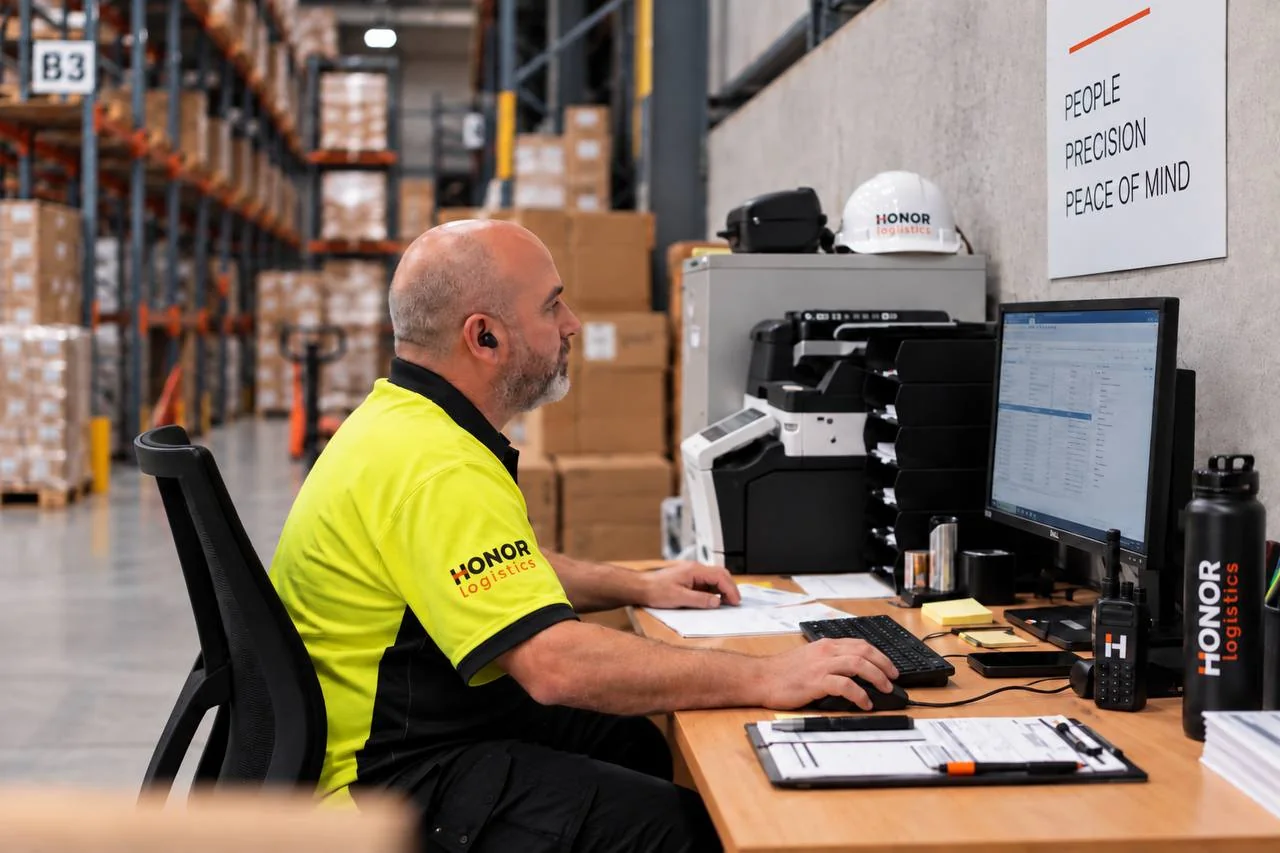 A Honor technician at a desk inside the warehouse, stock records on the monitor, a multifunction printer and a hard hat beside him, pallet racking behind.