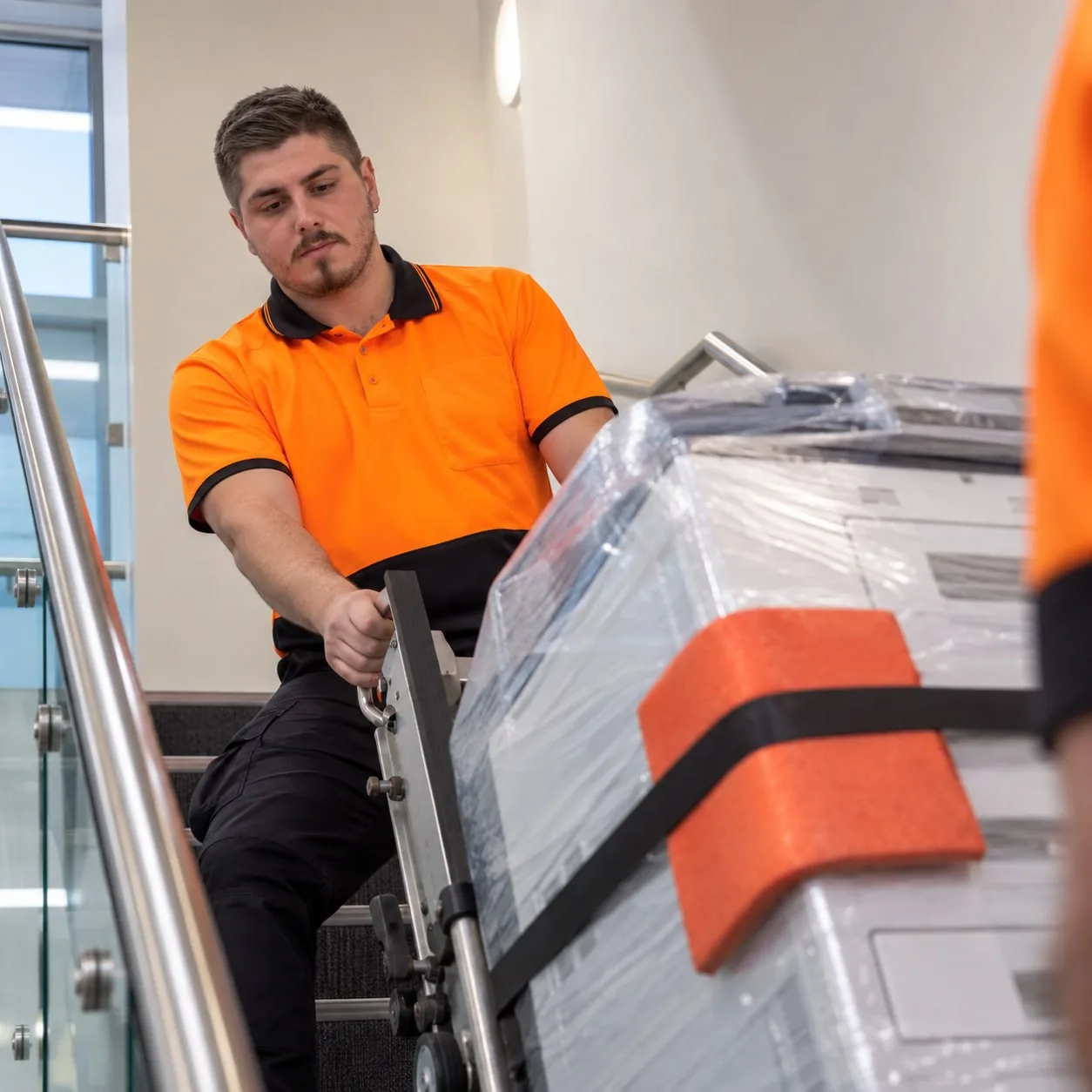 A Honor technician guiding a strapped, shrink-wrapped printer up a staircase on a stair-climber.
